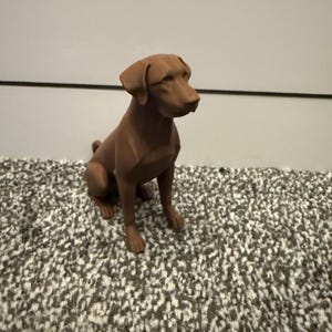 May include: A brown dog figurine in a seated position. The dog has a smooth, matte finish and is set against a neutral background. The figurine is a detailed representation of a dog breed.