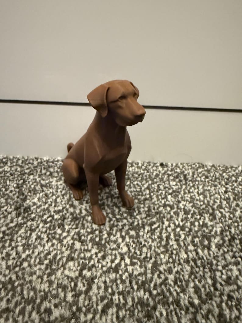 May include: A brown dog figurine in a seated position. The dog has a smooth, matte finish and is set against a neutral background. The figurine is a detailed representation of a dog breed.