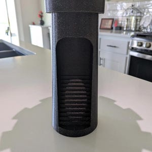 May include: A black, castle-shaped dice tower with a textured surface. The tower features a rectangular opening revealing a staircase inside. The top has crenellations, and the base is cylindrical.