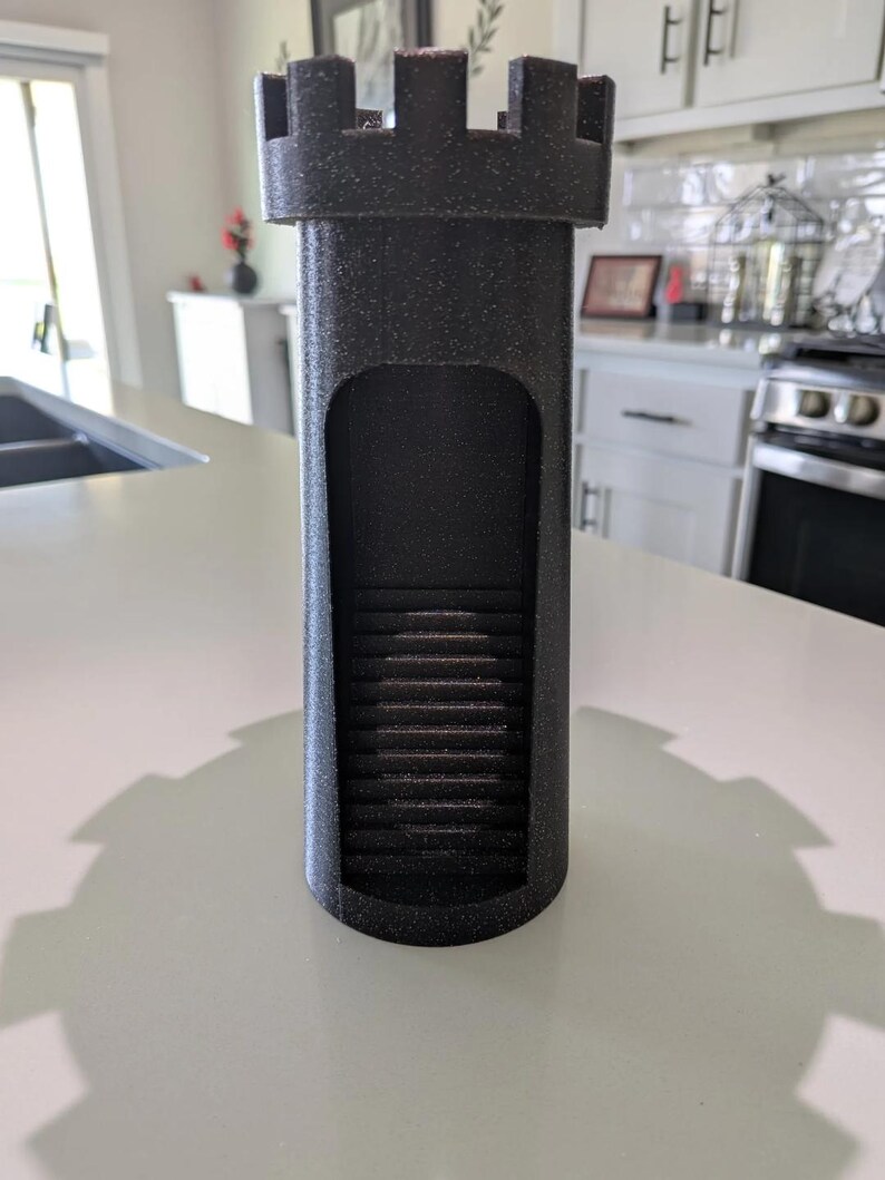 May include: A black, castle-shaped dice tower with a textured surface. The tower features a rectangular opening revealing a staircase inside. The top has crenellations, and the base is cylindrical.