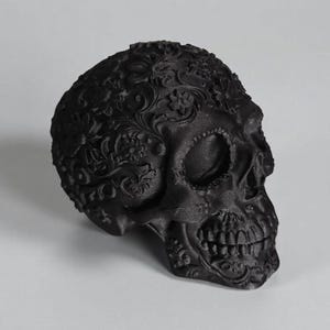 May include: A black decorative skull with intricate floral patterns carved across the surface. The skull features detailed teeth and eye sockets, creating a gothic and artistic aesthetic. The skull is a decorative item.