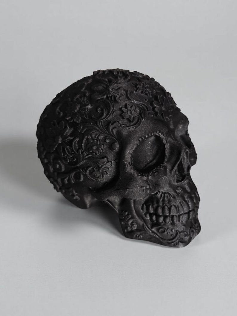 May include: A black decorative skull with intricate floral patterns carved across the surface. The skull features detailed teeth and eye sockets, creating a gothic and artistic aesthetic. The skull is a decorative item.