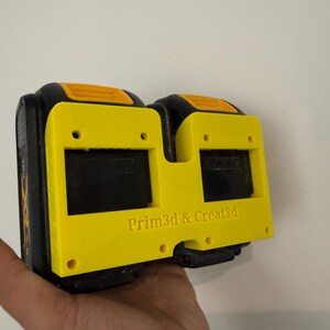 May include: A yellow and black 3D-printed battery holder with the text "Prim3d & Creat3d". The holder has two rectangular openings and is designed to hold two batteries. The holder is held in a hand.