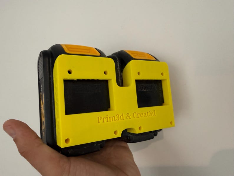May include: A yellow and black 3D-printed battery holder with the text "Prim3d & Creat3d". The holder has two rectangular openings and is designed to hold two batteries. The holder is held in a hand.