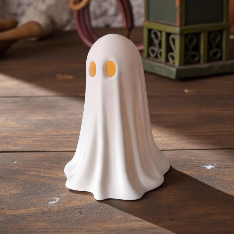 May include: A white ceramic ghost figurine with two orange eye holes. The ghost has a draped sheet-like form and is standing on a wooden surface. The figurine is a Halloween or spooky-themed decorative item.