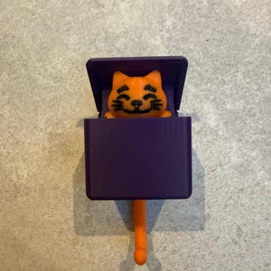 May include: A whimsical orange cat figurine peeks out from a purple box with a hinged lid. The cat has black facial features and is set against a neutral background. The box has an orange tail-like handle.