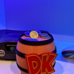 May include: A barrel-shaped coin bank with the letters "DK" in red and gold, reminiscent of the Donkey Kong video game series. Several coins rest on top of the bank. The bank is peach-colored with black trim.