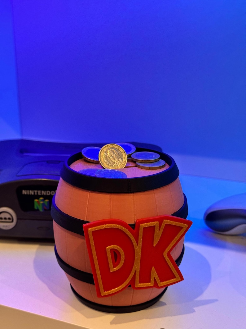 May include: A barrel-shaped coin bank with the letters "DK" in red and gold, reminiscent of the Donkey Kong video game series. Several coins rest on top of the bank. The bank is peach-colored with black trim.