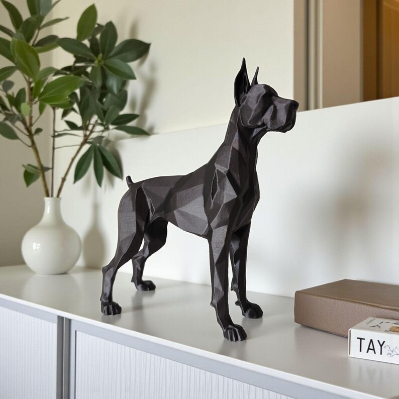 Puede incluir: Escultura de perro geom&eacute;trico negro con un dise&ntilde;o facetado. El perro est&aacute; de pie sobre sus cuatro patas, con las orejas erguidas. La escultura est&aacute; colocada sobre una superficie blanca, con una planta en un jarr&oacute;n blanco al fondo.