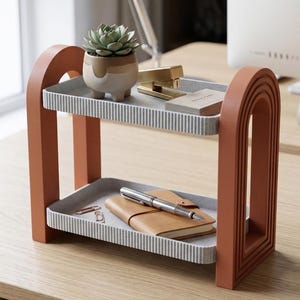 May include: A two-tiered, terracotta-colored desk organizer with a modern design. The shelves are gray with vertical striped edges. The top shelf holds a small succulent in a ceramic pot, a gold stapler, and business cards. The bottom shelf has a notebook and pen.