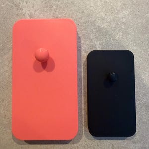 K&ouml;nnte beinhalten: Zwei rechteckige Flaschenverschl&uuml;sse. Einer ist korallenrosa mit einem runden Knopf, der andere ist schwarz mit einem dreieckigen Knopf. Beide haben abgerundete Ecken und liegen auf einer grauen Oberfl&auml;che.