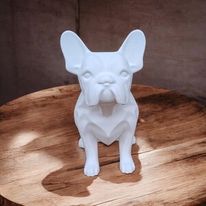May include: White ceramic figurine of a French bulldog sitting on a wooden surface. The figurine is stylized with geometric shapes.
