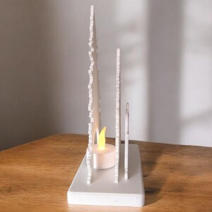 May include: A white decorative candle holder with a rectangular base and vertical, stylized tree-like cutouts. A small, battery-operated candle with a yellow flame is placed inside the holder. The candle holder is on a wooden surface.