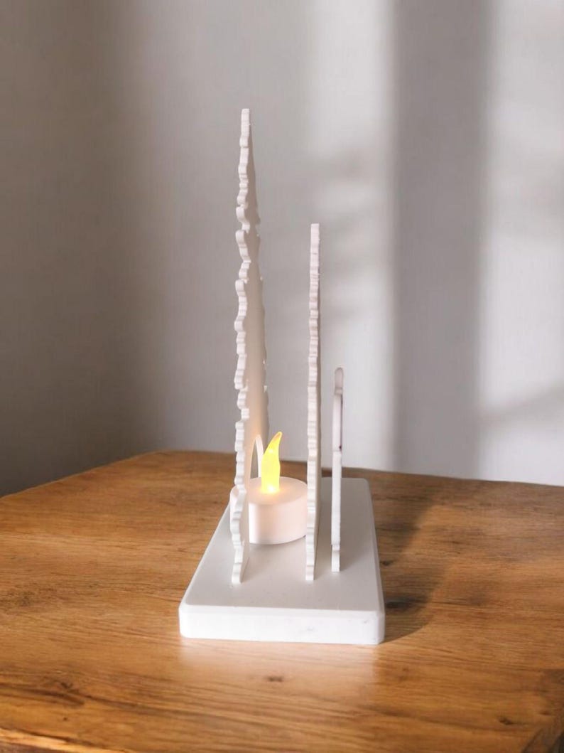 May include: A white decorative candle holder with a rectangular base and vertical, stylized tree-like cutouts. A small, battery-operated candle with a yellow flame is placed inside the holder. The candle holder is on a wooden surface.