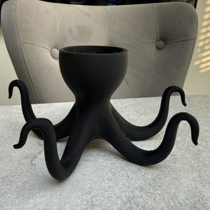 May include: A black octopus-shaped planter with a round top and eight curved tentacles. The planter is made of a matte material and sits on a light grey surface. The tentacles are evenly spaced and curl upwards.