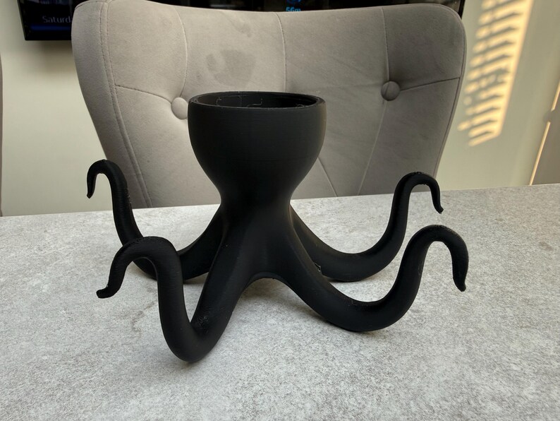 May include: A black octopus-shaped planter with a round top and eight curved tentacles. The planter is made of a matte material and sits on a light grey surface. The tentacles are evenly spaced and curl upwards.
