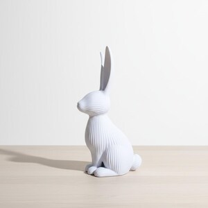 May include: A white ceramic rabbit figurine with long ears and a textured, ribbed design. The bunny is sitting upright on its hind legs, with its front paws resting on the surface. The figurine is a decorative item.