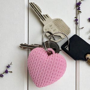 May include: A pink, knitted-effect heart-shaped keyring attached to a key ring with silver and black keys. The background is a white wooden surface, decorated with sprigs of purple flowers.