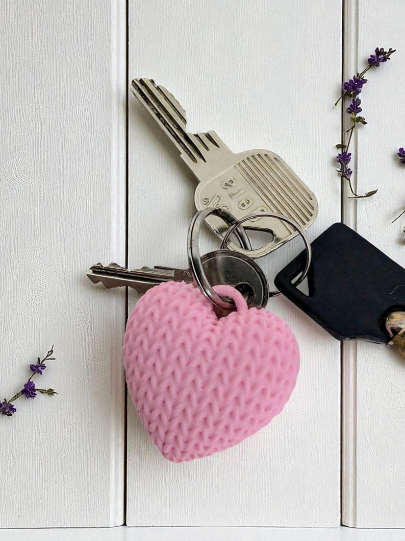 May include: A pink, knitted-effect heart-shaped keyring attached to a key ring with silver and black keys. The background is a white wooden surface, decorated with sprigs of purple flowers.