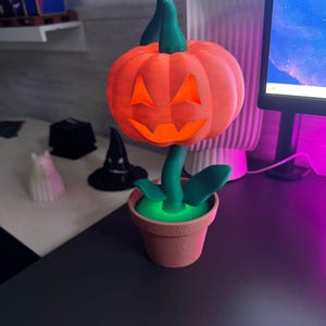 May include: A 3D-printed Halloween decoration featuring an orange pumpkin with a carved face, green stem and leaves, and a terracotta-colored pot. The pumpkin sits atop a green stem, and the pot has a glowing green interior.