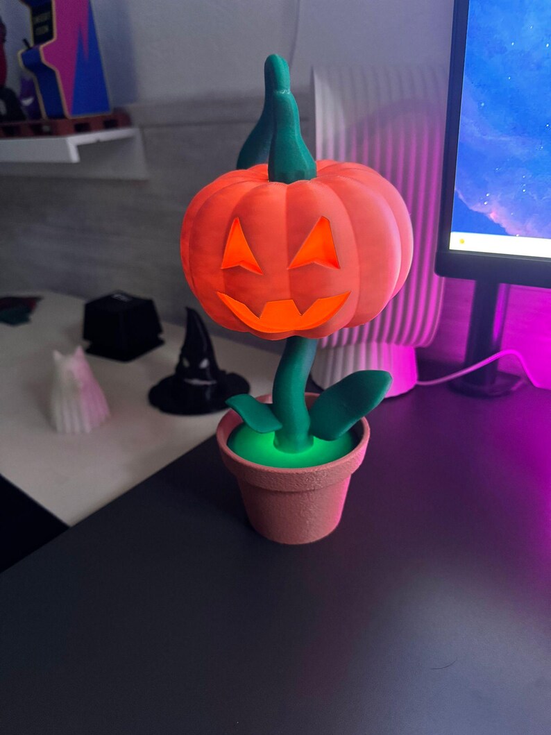 May include: A 3D-printed Halloween decoration featuring an orange pumpkin with a carved face, green stem and leaves, and a terracotta-colored pot. The pumpkin sits atop a green stem, and the pot has a glowing green interior.