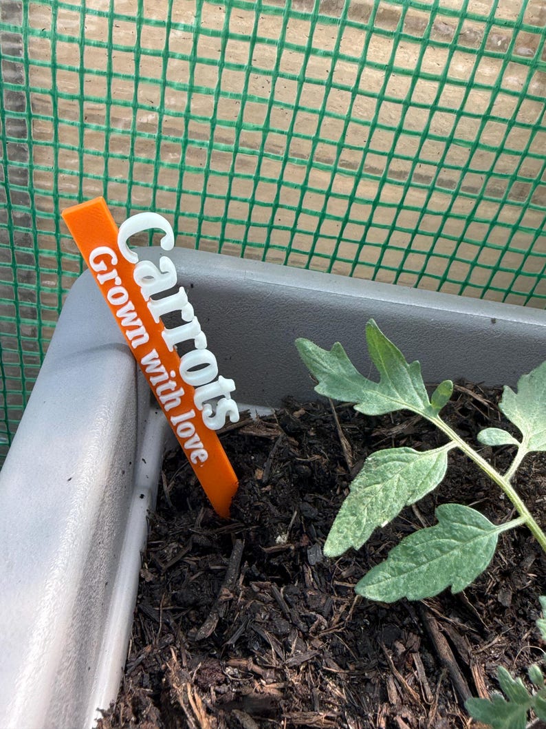 May include: An orange plant marker with the word "Carrots" in white, and the phrase "Grown with love". The marker is placed in a gray planter with dark soil, next to a small green plant. A green mesh fence is in the background.