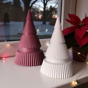 May include: Two decorative Christmas tree figurines. One is a dusty rose color, the other is white. Both have a ribbed design and are placed on a white surface near a window. A red poinsettia plant is in the background.