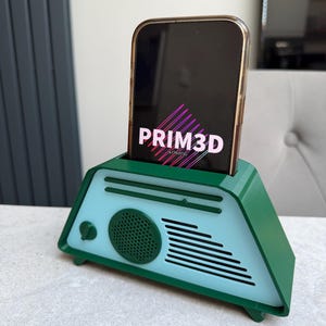 May include: A green and light blue 3D-printed phone stand shaped like a retro radio. The phone, displaying "PRIM3D", is held upright in the stand. The stand has a speaker and a dial.