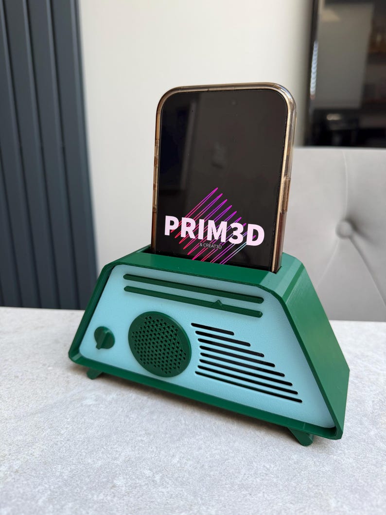 May include: A green and light blue 3D-printed phone stand shaped like a retro radio. The phone, displaying "PRIM3D", is held upright in the stand. The stand has a speaker and a dial.