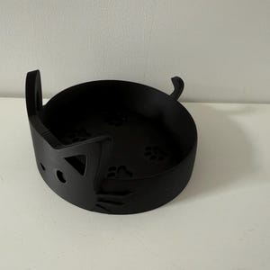 Puede incluir: Un cuenco negro con forma de gato con recortes de huellas de patas en la base. El cuenco tiene forma redondeada con un dise&ntilde;o de cara de gato en el lateral, incluyendo asas en forma de orejas. El cuenco est&aacute; hecho de un material negro mate.
