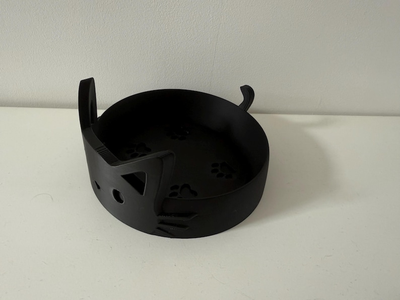 Puede incluir: Un cuenco negro con forma de gato con recortes de huellas de patas en la base. El cuenco tiene forma redondeada con un dise&ntilde;o de cara de gato en el lateral, incluyendo asas en forma de orejas. El cuenco est&aacute; hecho de un material negro mate.