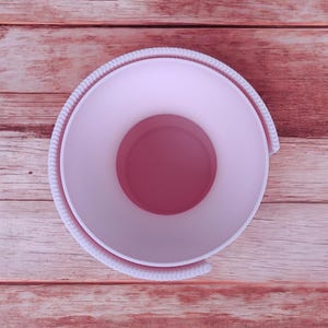 Puede incluir: Vista a&eacute;rea de un recipiente circular rosa claro con un interior rosa m&aacute;s oscuro. El recipiente tiene un borde texturizado en forma de cuerda. El fondo es una superficie de madera con un tono marr&oacute;n rojizo.