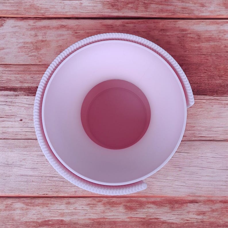 Puede incluir: Vista a&eacute;rea de un recipiente circular rosa claro con un interior rosa m&aacute;s oscuro. El recipiente tiene un borde texturizado en forma de cuerda. El fondo es una superficie de madera con un tono marr&oacute;n rojizo.