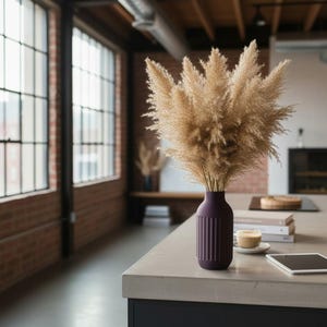 May include: A decorative arrangement of dried pampas grass in a dark purple vase. The vase has vertical ribbed detailing. The arrangement sits on a light gray countertop with a cup of coffee and a tablet nearby. The room has large windows.