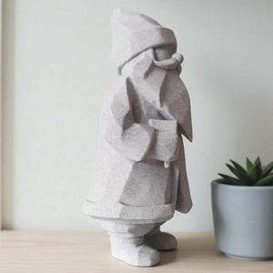 K&ouml;nnte beinhalten: Eine hellgraue geometrische Gartenzwerg-Statue. Der Zwerg tr&auml;gt einen Hut und einen Mantel und hat einen langen Bart. Die Statue steht auf einem Holzregal neben einer kleinen Topfpflanze. Der Zwerg ist ein dekorativer Artikel.