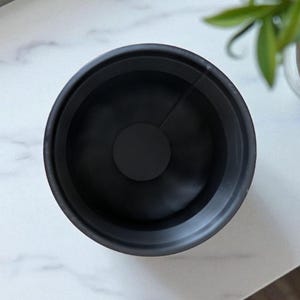 May include: A top-down view of a black, circular object with a central disc. The object has a ridged texture on the inside and is set against a white marble surface. The object appears to be a lens hood or similar photographic accessory.