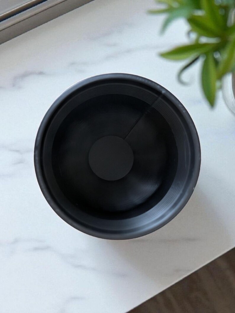 May include: A top-down view of a black, circular object with a central disc. The object has a ridged texture on the inside and is set against a white marble surface. The object appears to be a lens hood or similar photographic accessory.