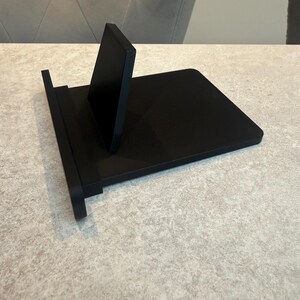May include: A black phone stand with a minimalist design. The stand features a rectangular base and a vertical support. It is designed to hold a phone or tablet. The stand is constructed from a solid, matte black material.
