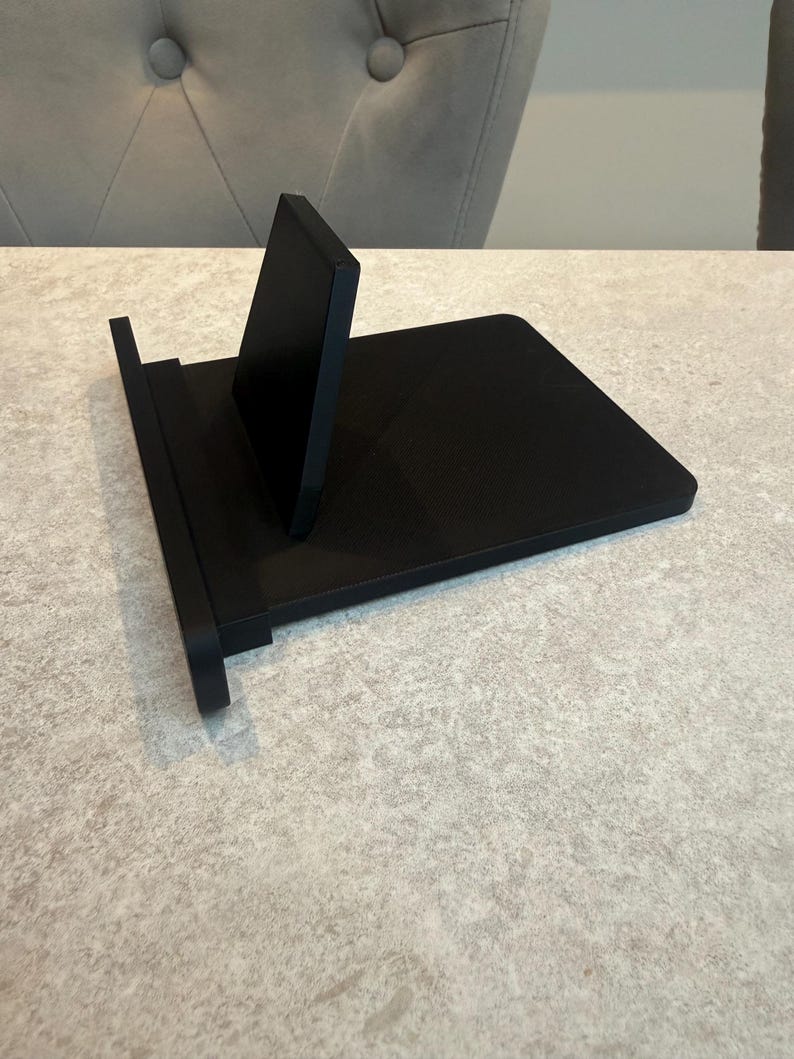 May include: A black phone stand with a minimalist design. The stand features a rectangular base and a vertical support. It is designed to hold a phone or tablet. The stand is constructed from a solid, matte black material.