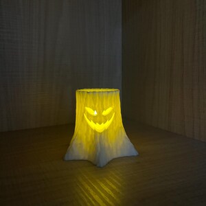 Puede incluir: Un portavelas tem&aacute;tico de Halloween con forma de toc&oacute;n de &aacute;rbol, con una cara de linterna tallada. El portavelas es blanco y amarillo y brilla desde el interior. Se coloca sobre una superficie de madera, creando un ambiente c&aacute;lido.