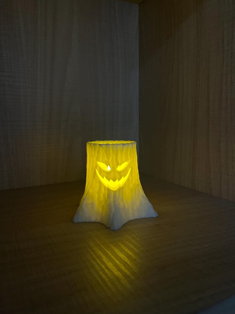 Puede incluir: Un portavelas tem&aacute;tico de Halloween con forma de toc&oacute;n de &aacute;rbol, con una cara de linterna tallada. El portavelas es blanco y amarillo y brilla desde el interior. Se coloca sobre una superficie de madera, creando un ambiente c&aacute;lido.