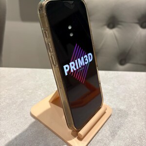 May include: A peach-colored phone stand holding a smartphone with a clear case. The phone displays the text "PRIM3D" in a stylized font with a pink and purple gradient. The stand is made of a matte material.