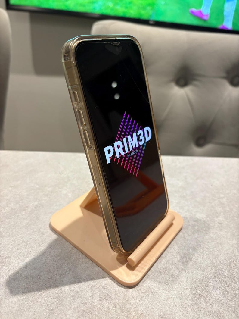 May include: A peach-colored phone stand holding a smartphone with a clear case. The phone displays the text "PRIM3D" in a stylized font with a pink and purple gradient. The stand is made of a matte material.