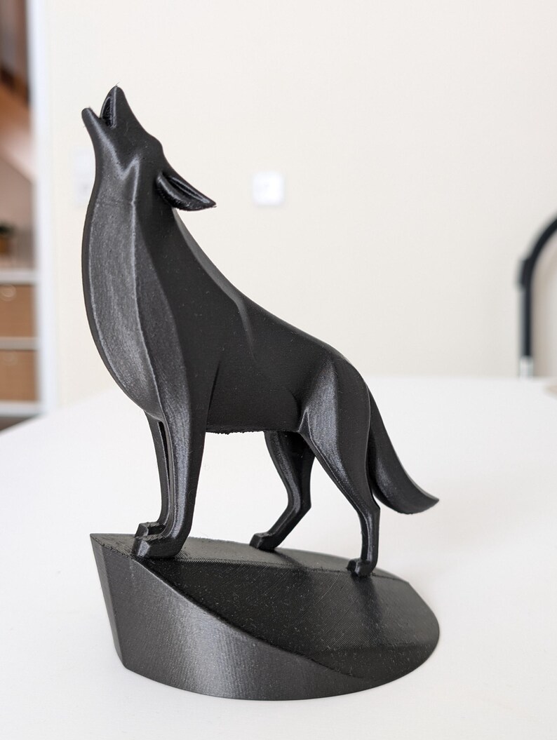 May include: A black, geometric wolf sculpture in a howling pose. The wolf is standing on a small, angled base. The sculpture is made of a matte black material and is set against a neutral background.