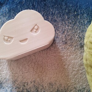 May include: A light pink cloud-shaped object with a carved face featuring angry eyes and a smiling mouth. The object is resting on a textured surface with blue and white hues, with a portion of a green plush item visible.