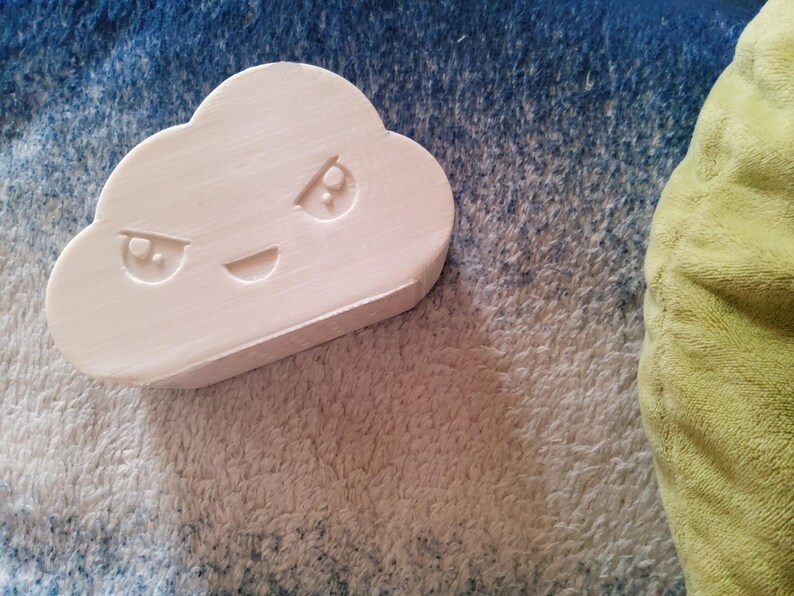 May include: A light pink cloud-shaped object with a carved face featuring angry eyes and a smiling mouth. The object is resting on a textured surface with blue and white hues, with a portion of a green plush item visible.