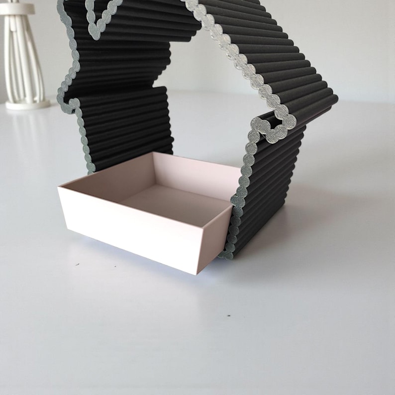 May include: A modern, black, house-shaped decorative shelf with a light pink rectangular tray. The shelf is constructed from layered, ridged material. The design is minimalist and geometric, suitable for displaying small items.
