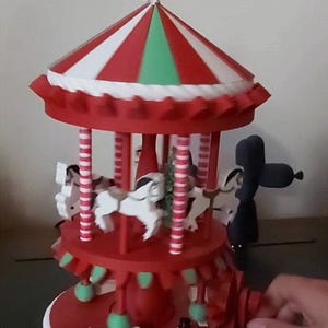 Op de afbeelding: Een rood en witte carrousel met een sneeuwvlok bovenop. De carrousel heeft witte paarden en is versierd met groene en witte accenten. De basis is rood met witte rand, en een kleine sneeuwman is zichtbaar.