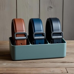 May include: Three leather belts in a light blue belt organizer. The belts are brown, dark blue, and navy blue. The organizer has three compartments.