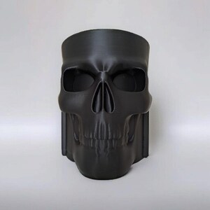 May include: A black skull-shaped planter with a wide opening. The planter has detailed facial features, including eye sockets, a nose cavity, and a jawline. The skull is made of a matte black material and is suitable for indoor or outdoor use.